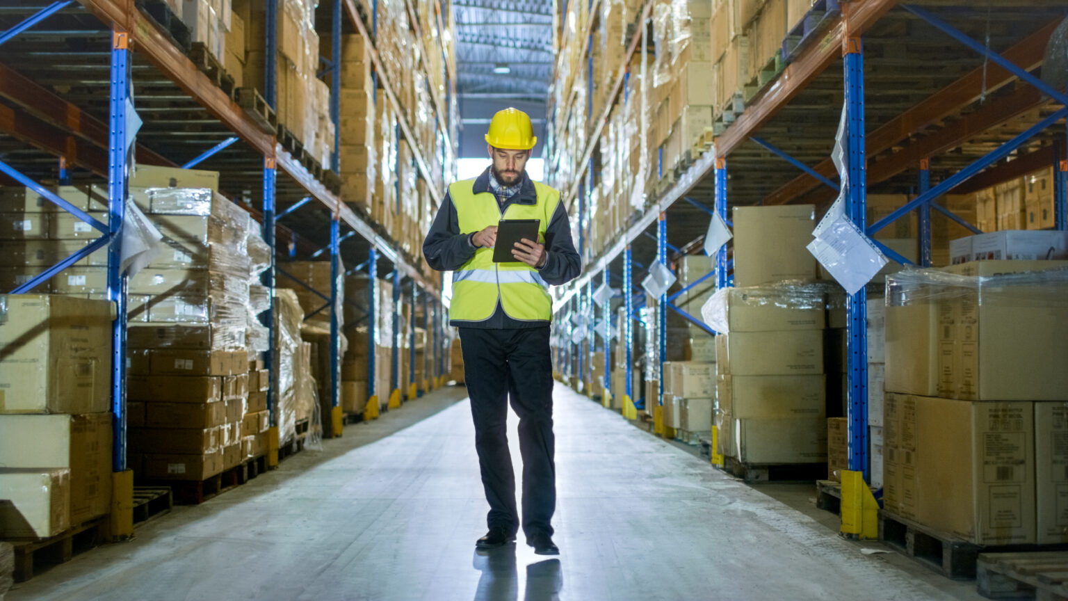 Auditor Wearing Hard Hat with Tablet Computer Counts Merchandise in Warehouse. He Walks Through Rows of Storage Racks with Merchandise.