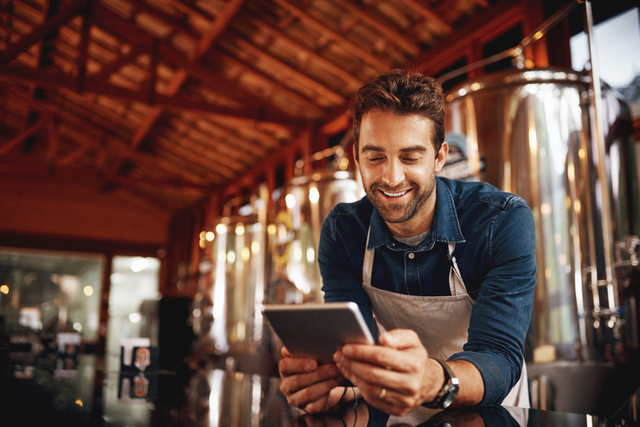 Shot of a cheerful young barman browsing on a digital tablet while patiently waiting at the bar for customers inside of a beer brewery during the day