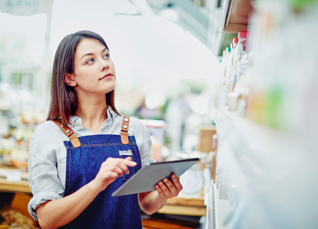 Young deli owner holding digital tablet while standing in store. Beautiful saleswoman is examining food on shelves in shop. She is wearing denim overalls.