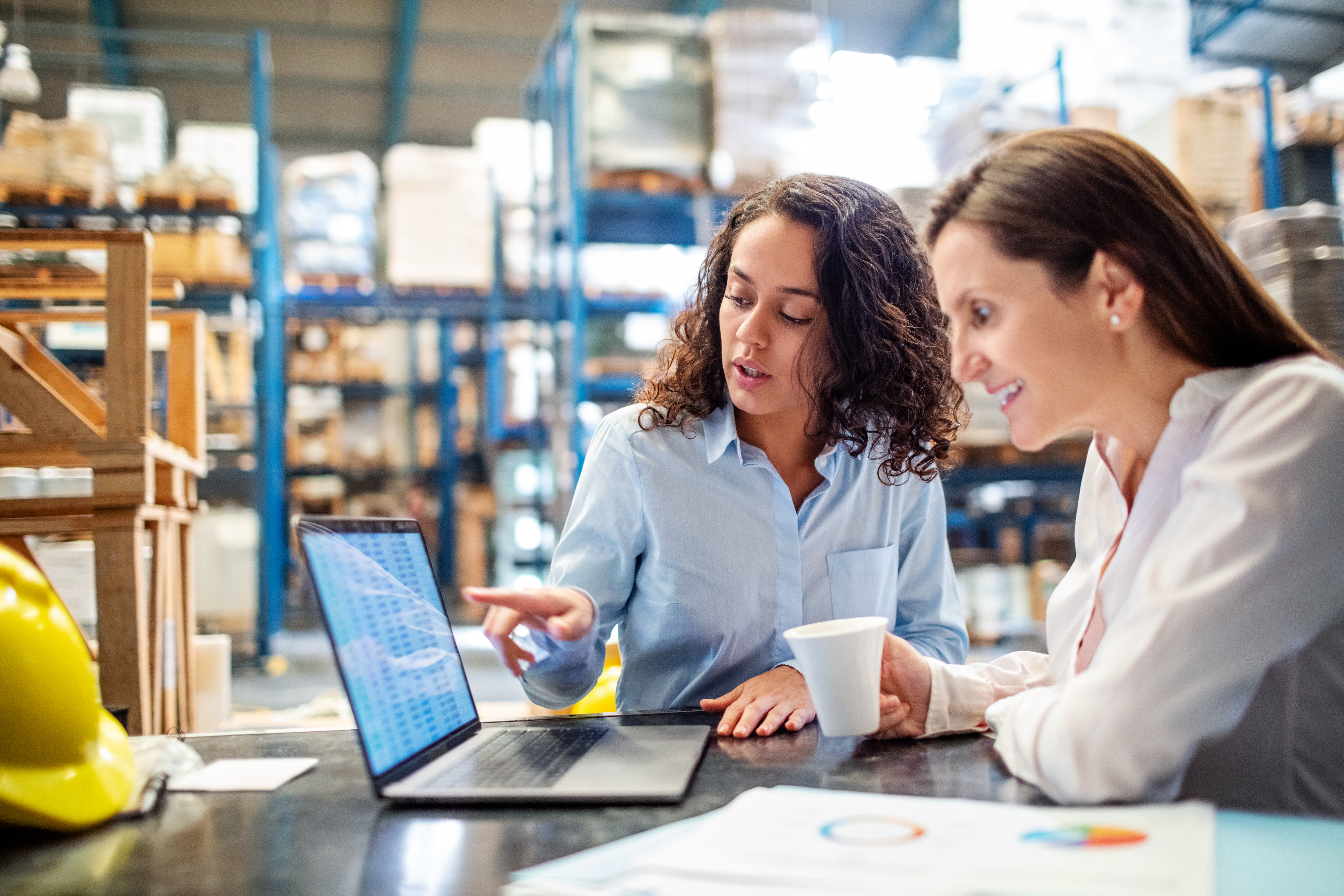 Young woman showing something on laptop to manager in factory warehouse. Two business women working together on laptop, checking the inventory.