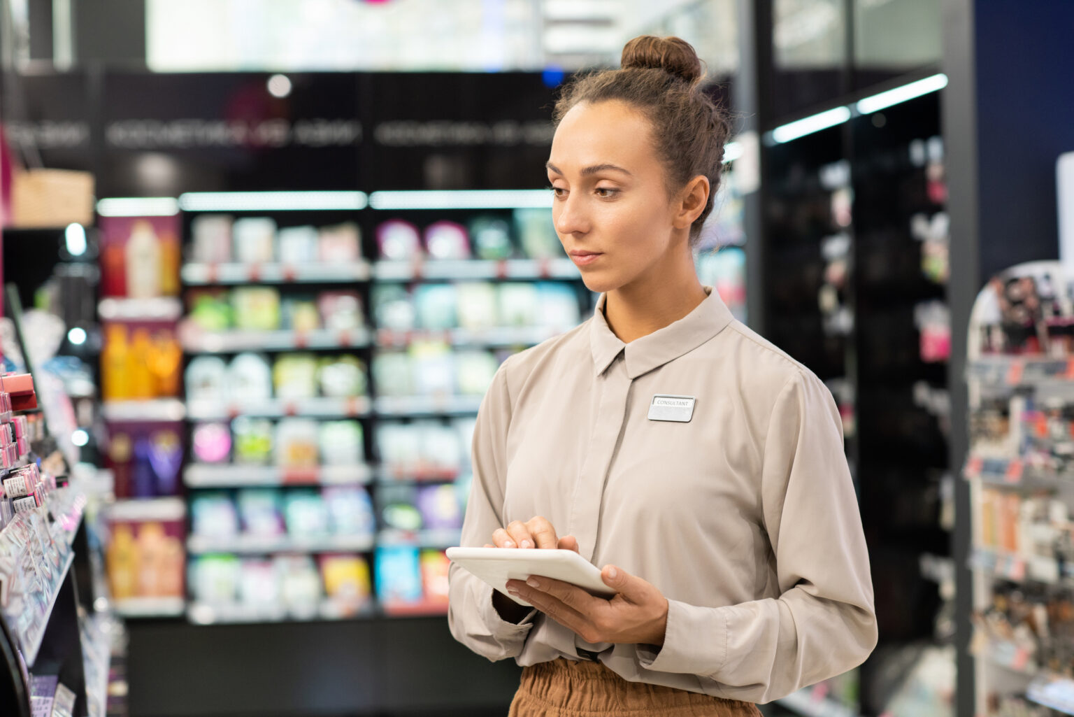 Young serious female consultant with touchpad entering data while looking at large display with various beauty products during work