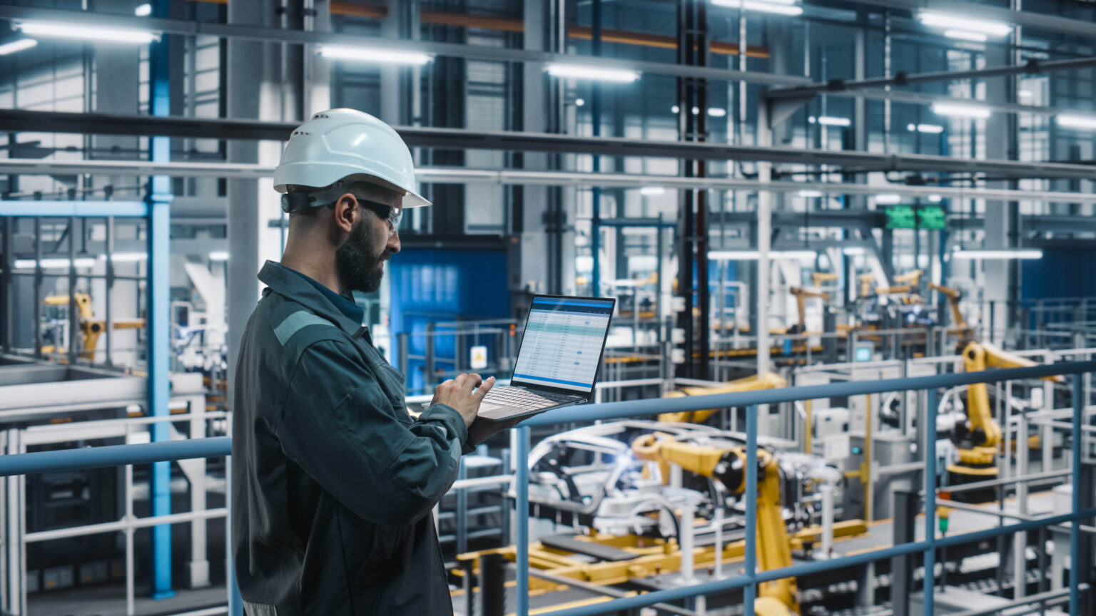 Car Factory Engineer in Work Uniform Using Laptop Computer with Spreadsheet Software. Working with Software at Automotive Industrial Manufacturing Facility Dedicated for Vehicle Production.