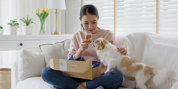 Young happy asia people girl smile enjoy with cute dog unbox snack food post mail sit relax at home comfort sofa couch in omni channel fast send parcel via online sale pet shop store e-commerce order.